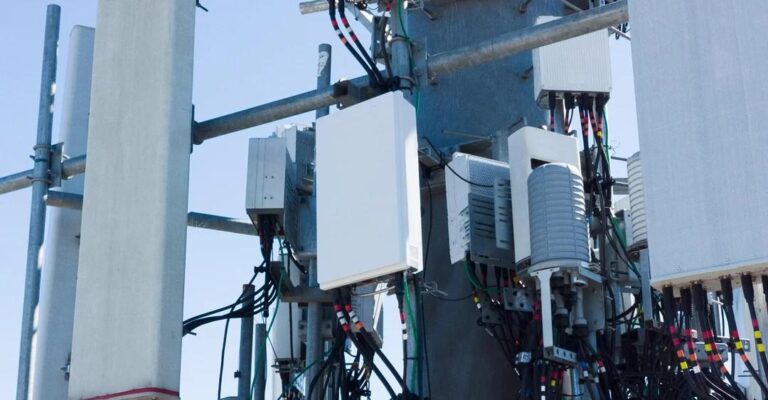 Primer plano de una torre de telecomunicaciones con múltiples antenas y equipos de 5G instalados, mostrando cables y cajas blancas sobre un fondo de cielo azul claro